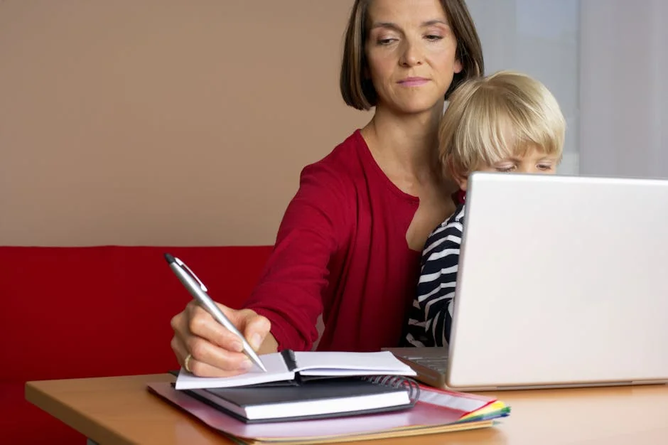 A woman multitasks with a child on her lap while working on a laptop at home.